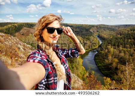 Young blonde smiling, woman tourist  on a cliff taking selfie picture on background of autumn landscape with the river Berd. Russia, Siberia, Salair