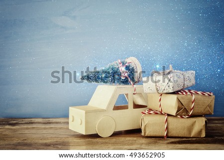 Wooden car carrying a christmas tree next to christmas gifts. Glitter overlay