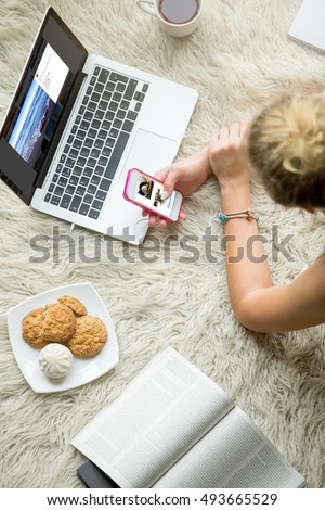 Beautiful young woman lying on white carpet with cup of tea and cookies, browsing social media, looking at pictures on smartphone and laptop, studying and relaxing at home. Close-up top view image