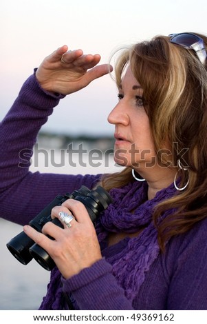 A woman looking at something off in the distance with binoculars in her hands while at the seashore.