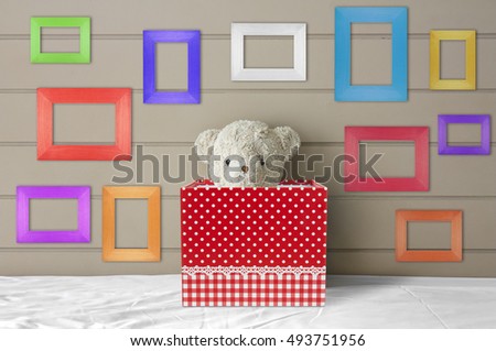 teddy bear doll in red gift box on the white bed with blank multi-color or fancy picture frame at the headboard and brown wall background for gift and surprise