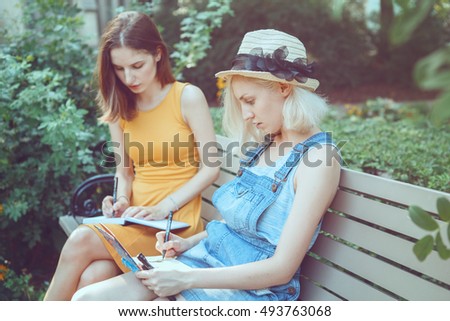 Portrait of two white Caucasian young girls hipster students friends outside in park on summer day sitting on bench together, drawing, sketching, best friends forever, toned with filters