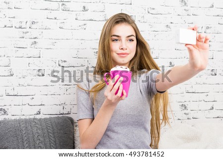 Cheerful pretty blonde teenage girl at home, holding pink coffee mug, taking a selfie with action camera, smiling. Brick wall background, no retouch, no filter.