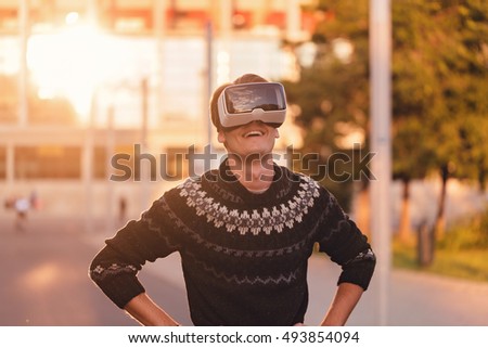 Amused young man with one hand in his pocket wearing a pair of VR glasses in a cool sweater and trendy outfit excited by augmented reality sunlit by an amazing sunset reflection from background