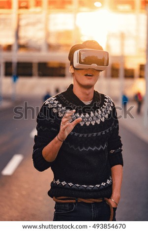 Amazed young man with one hand touching the air wearing a pair of VR glasses in a cool sweater and trendy outfit impressed by augmented reality sunlit by an amazing sunset reflection in the background