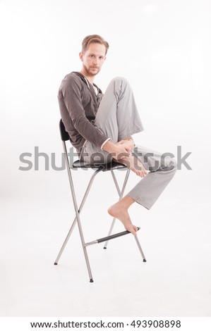 Young adult male sitting on a bar chair barefoot isolated on white background.