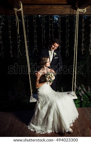 Wedding picture of a happy couple, she is sitting on the swings he is standing beside her holding her shoulders smiling happily at her