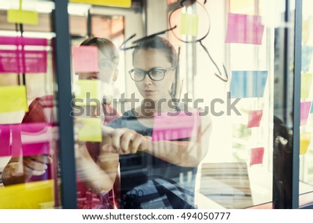 Two business women working together on wall glass with post it stickers. Modern startup office Royalty-Free Stock Photo #494050777