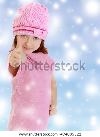 Cheerful little girl in a pink knitted dress and hat, shows hand gesture all right, thumb to the top. Blue Christmas festive background with white snowflakes.