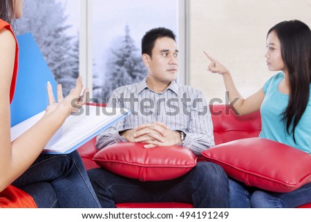 Portrait of husband and wife sitting on the red couch and arguing problem while psychiatrist talking, winter on the window