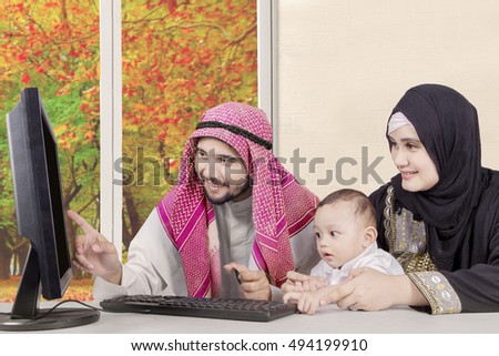 Image of Arabian family using computer and holding credit card while smiling and looking at computer at home with autumn background on the window