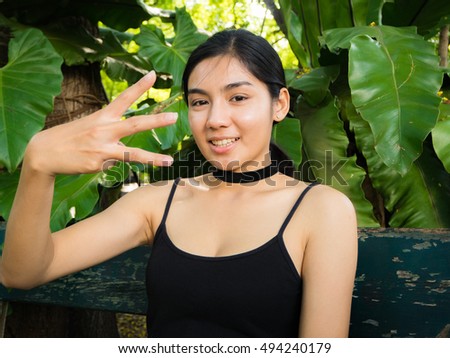 Asian woman make a "E" from her hand, sitting on chesterfield, black hair, black vest, green leaves background, portrait