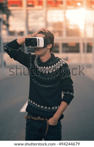 Amazed young man in soft focus with one hand in his pocket wearing a pair of VR glasses in a cool sweater and trendy outfit excited by augmented reality sunlit by an sunset reflection