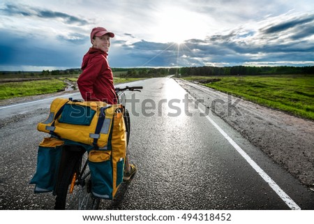 Woman riding loaded bicycle on the wet asphalt road with clouds on the background