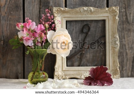 Floral scene with beautiful cream rose, pink flower,vinous leaves,pink sweet peas in vintage green glass vase, petals of roses, wooden rectangular golden frame with scissors on dark wooden background 