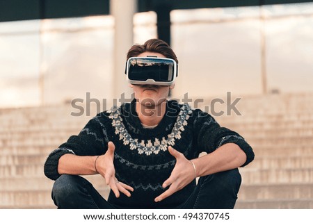 Angry young man wearing virtual reality VR glasses in a cool sweater and trendy outfit disappointed by augmented reality sunlit by an amazing sunset reflection in the background