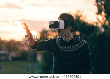 curious young man wearing a pair of VR glasses with hands touching virtual reality in a cool sweater and trendy outfit amazed by augmented reality sunlit by an amazing LOW LIGHT sunset soft focus