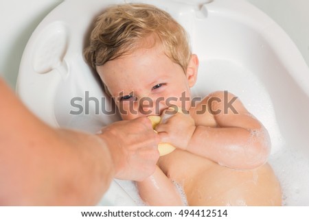 Little pretty wet baby boy in bath room sitting and playing with mother's hand and sponge on white background, horizontal picture.