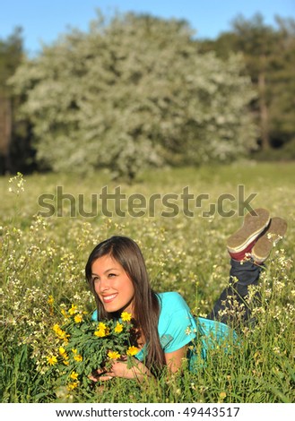 Beautiful young blonde woman lying in blooming meadow in spring, bunch of yellow flowers in hand, smiling,  blue sky and trees in background; shallow depth of field