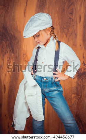 Children's emotions. Artistic little girl in jeans with suspenders, white shirt and a cap on the wooden background