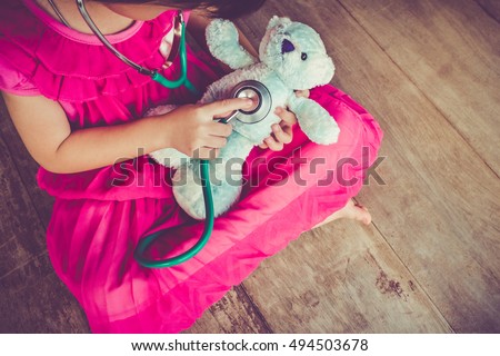 Top view of child playing doctor or nurse with plush toy bear with bright sunlight at home. Happy girl listens a stethoscope to toy. Playful girl role playing. Vintage tone effect.
