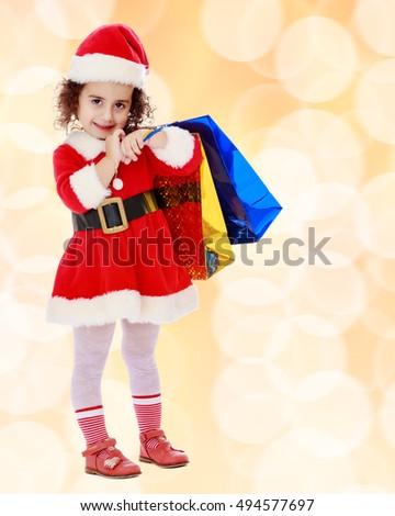 Caucasian little curly-haired girl in a coat and hat of Santa Claus carries on his shoulder a colorful shopping bags.Winter brown abstract background with white snowflakes.