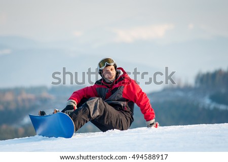 Smiling snowboarder wearing helmet, red jacket, gloves and pants sitting on snowy slope on top of a mountain, with an astonishing view on hills