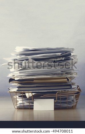 A wire office tray with blank label, piled up with papers and folders, undersaturated in drab hues for dreary, dystopian feel.