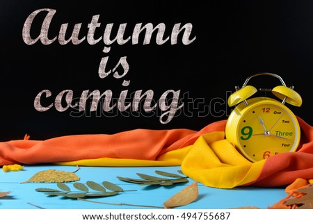 Alarm clock near a black school board, autumn leaves, scarf and an inscription Autumn is coming