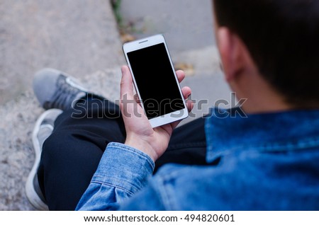 Close up white mobile phone using in hand a young hipster business man  sitting and looking at phone in denim jeans blue shirt pants black shoes gray running shoes of a stone slab paving slabs Royalty-Free Stock Photo #494820601
