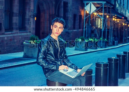 Asian American college student studies in New York. Wearing black leather jacket, young guy sits on metal pillar on narrow vintage street, looks away, thinks, works on laptop compute. Filtered effect.