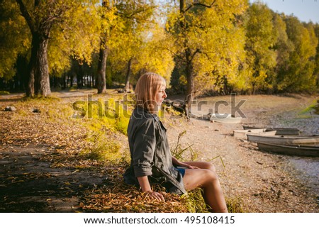 pretty blonde girl in sneakers, nylon stockings and coat, sits on  banks of river, on yellow leaves, enjoying warmth of autumn sun rays