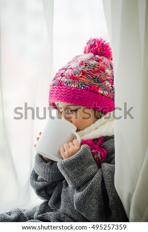 Cute little girl in fashionable winter clothes with a knitted hat and warm sweater. Holding in her hand a white Cup of tea.