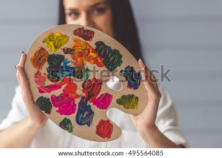 Attractive young female artist is holding a color palette, looking at camera and smiling while standing in her studio