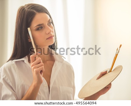 Attractive young female artist is holding paint brushes and a palette, looking away and thinking while standing in her studio