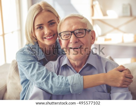 Handsome old man and beautiful young girl are hugging, looking at camera and smiling while sitting on couch at home Royalty-Free Stock Photo #495577102