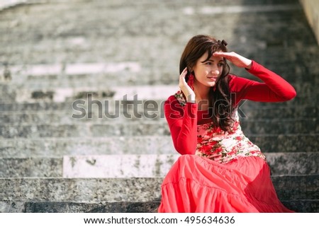 Woman in beautiful red dress with roses on it. She sits on stairs, keeps her hand to protect her eyes against sun and looking somewhere far. Fashion or beauty kind of photography. Place for your text.