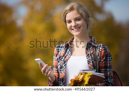 Portrait of a young attractive student outsides, holding a mobile phone in one hand, yellow leaves in another, yellow background. Fall time. Back to school concept photo, looking at the camera
