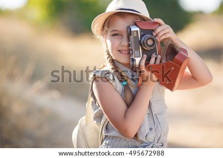 small child girl holding camera and taking photo of sunset landscape, standing on hill with background of mountains and sea, traveling photographer. girl with backpack, suitcase, summer hat