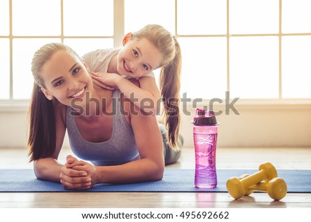 Beautiful young woman and charming little girl are looking at camera and smiling while lying on yoga mat in fitness hall Royalty-Free Stock Photo #495692662