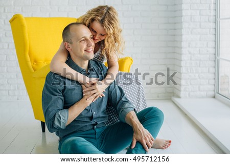 Happy couple in love in white room. Woman with curly hair sitting in yellow armchair hugging her boyfriend in green pants sitting on the floor. Laughing and smiling family of two.
