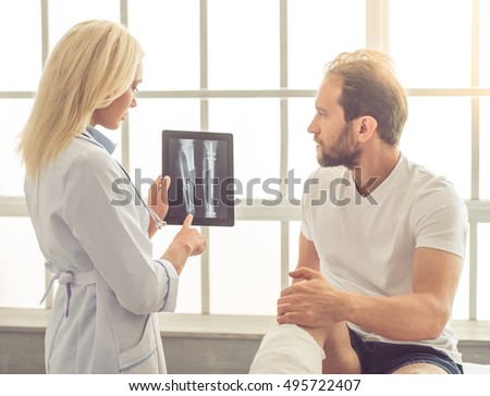 Beautiful female medical doctor is talking to handsome patient with broken leg and showing him X-ray picture on tablet