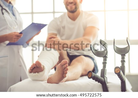 Cropped image of beautiful female medical doctor listening to handsome patient with broken leg and making notes while working in her office Royalty-Free Stock Photo #495722575