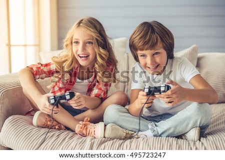 Pretty little girl and boy are playing game console and laughing while sitting on sofa at home