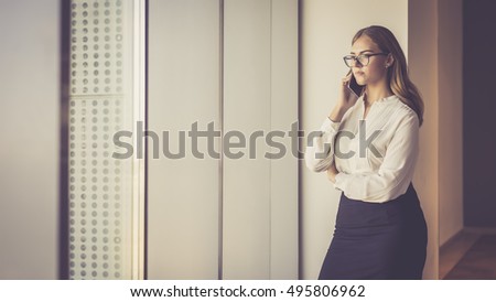 Young businesswoman standing near window and talking on cellphone.Girl in a black skirt and white blouse, standing at the window and talking on mobile phone. Young woman using digital gadget.