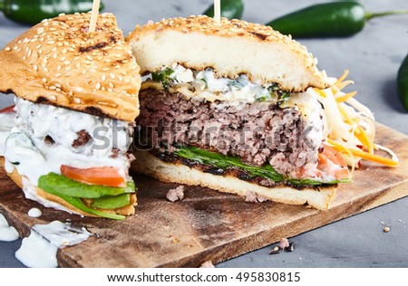 Gourmet homemade burger with beef, fried spicy jalapenos, fresh spinach, sliced tomatoes, cheese, grilled bun and sauce of yogurt and blue cheese. With coleslaw on wooden board. Jalapenos behind