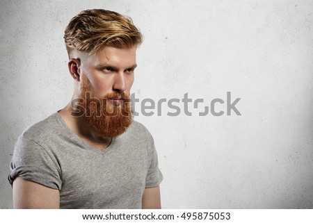 Portrait of courageous and fashionable male model with long trendy beard and undercut hairstyle. Caucasian blond man in grey T-shirt looking sullenly ahead of him. Indoors shot on white background. Royalty-Free Stock Photo #495875053