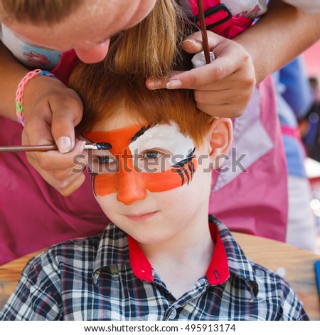 Artist's hand draws face painting to little boy. Child with funny face art. Painter makes tiger eyes on boy's face. Children holiday, event, birthday party, entertainment.