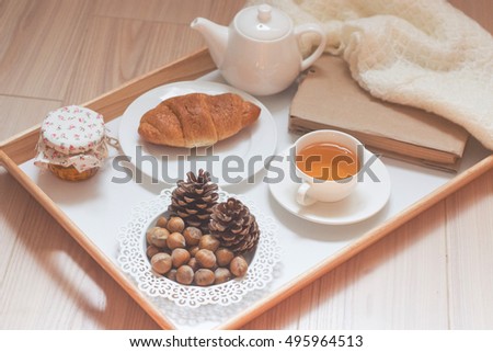 A cup of hot tea, a croissant on a white plate, a book, a white teapot, a knitted blanket, nuts and a jar of honey on a wooden tray.