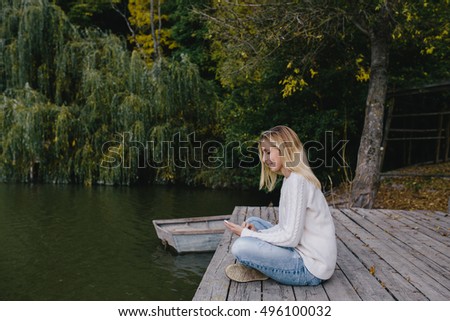 young blonde woman in a white sweater and blue jeans sitting on a wooden pier on the background of autumn forest, lake and using smart phone. woman sending a text message from her cell phone outdoors